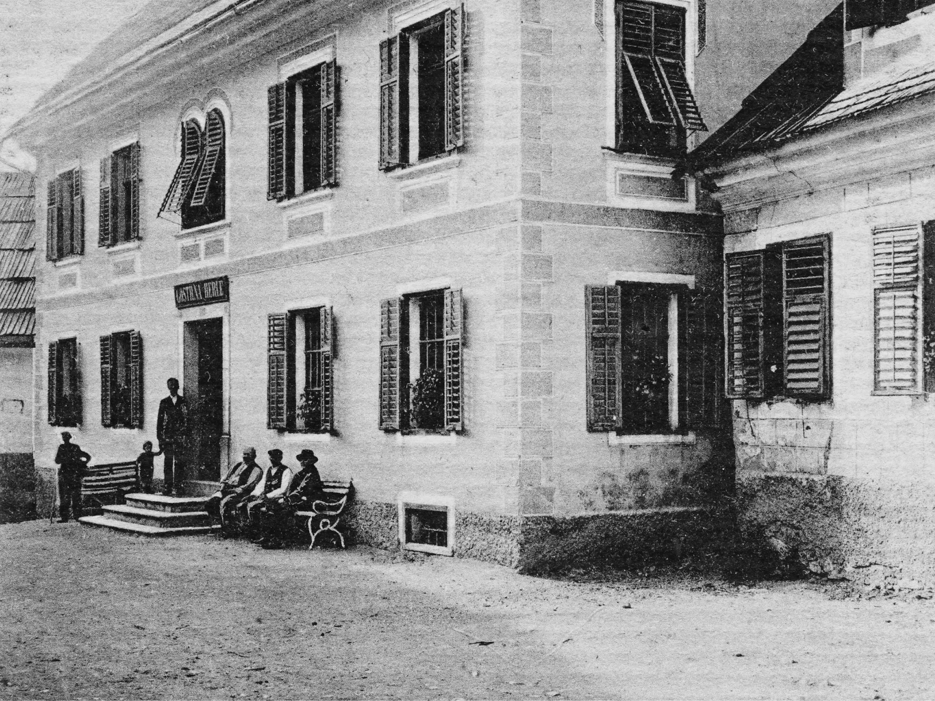 Starejša črno-bela fotografija zunanjosti gostilne, pred njo na klopci sedijo in zraven stojijo moški